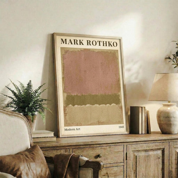 May include: Framed art print with abstract horizontal color blocks in muted pink, olive green, and cream tones, displayed on rustic wooden sideboard alongside potted fern, ceramic vase, and stacked books in neutral-toned living room setting with white walls and upholstered furniture.