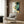 May include: Modern living room with beige upholstered armchair and geometric throw pillow. Large framed art print on wood-paneled wall features flowing abstract shapes in green, gold, and navy with Japanese calligraphy text. Golden floor lamp and dark round side table with open book complete the sophisticated interior setting.