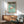 May include: Japanese flamingo poster with pink bird on turquoise background, displayed in modern living room with mid-century wooden sideboard, sage green armchair, white ceramic lamp, and white wall with natural sunlight shadows. Poster shows vintage exhibition design with Japanese text and appears framed in light wood frame.