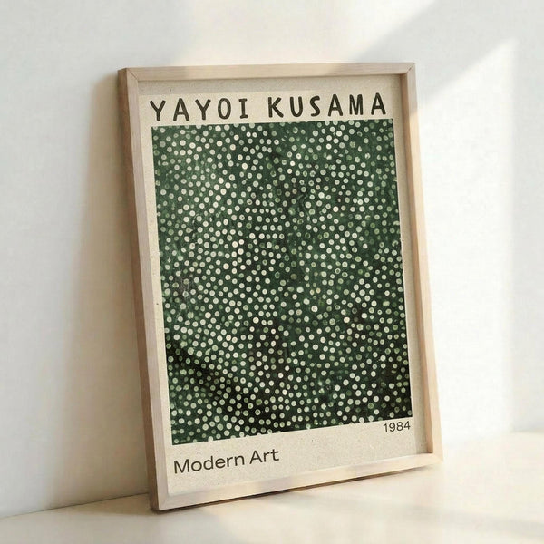 May include: Framed art print featuring Yayoi Kusama's signature polka dot pattern in varying shades of green with white dots creating an infinity net design. The minimalist poster has "YAYOI KUSAMA" at top, "1984" and "Modern Art" at bottom on cream background. Displayed in light wood frame leaning against white wall with natural lighting casting soft shadows.