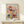 May include: Framed art print displaying colorful stylized flowers and botanical elements in red, yellow, blue, and pink with dotted patterns and geometric shapes, mounted in light wood frame on white shelf next to small wrapped gift with twine and greenery sprig against cream wall