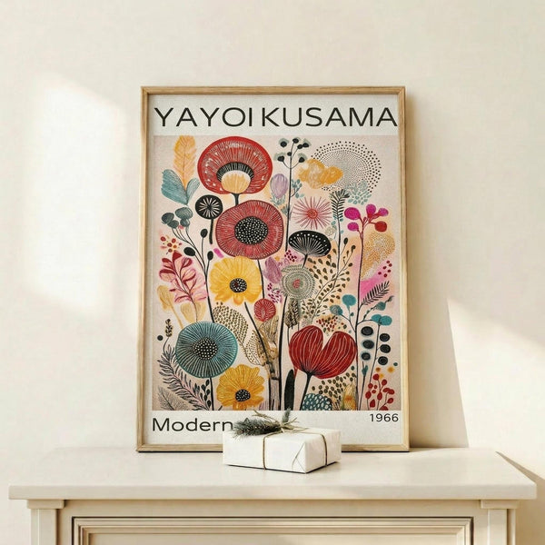 May include: Framed art print displaying colorful stylized flowers and botanical elements in red, yellow, blue, and pink with dotted patterns and geometric shapes, mounted in light wood frame on white shelf next to small wrapped gift with twine and greenery sprig against cream wall