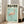 May include: Framed Japanese exhibition poster featuring pink flamingo on mint green background, displayed on light wooden floor against white wall beside beige upholstered chair. Poster shows Japanese text at top, dates 2007年5月3日-7月14日, and curator information at bottom. Natural lighting creates soft shadows in bright, minimalist room setting.
