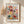 May include: Framed botanical art print in light wood frame hanging on beige wall, featuring colorful stylized flowers and plants in red, yellow, teal, and pink with geometric patterns and dotted details. Dried pampas grass stems visible on left side. Soft natural lighting creates gentle shadows on wall.