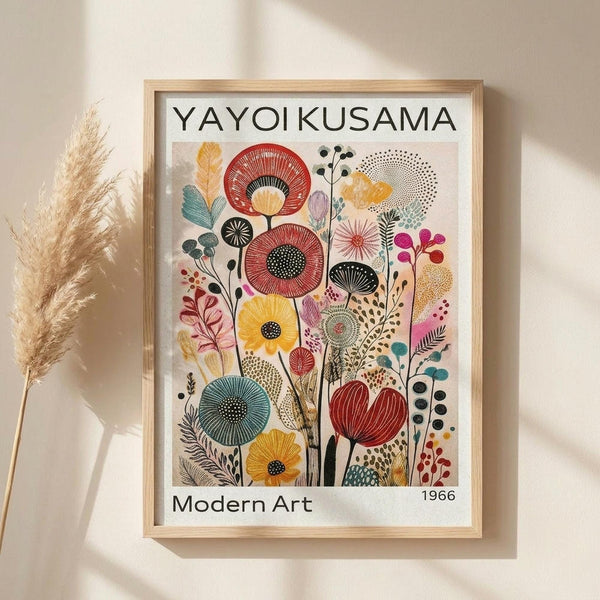 May include: Framed botanical art print in light wood frame hanging on beige wall, featuring colorful stylized flowers and plants in red, yellow, teal, and pink with geometric patterns and dotted details. Dried pampas grass stems visible on left side. Soft natural lighting creates gentle shadows on wall.