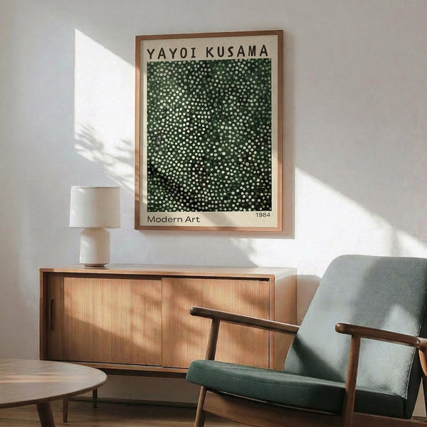 May include: Green and white dotted pattern artwork in wooden frame hanging above mid-century modern wooden credenza with white ceramic lamp. Sage green upholstered armchair with wooden arms sits nearby on wooden floor. Natural sunlight creates shadows on white wall in minimalist living room setting.