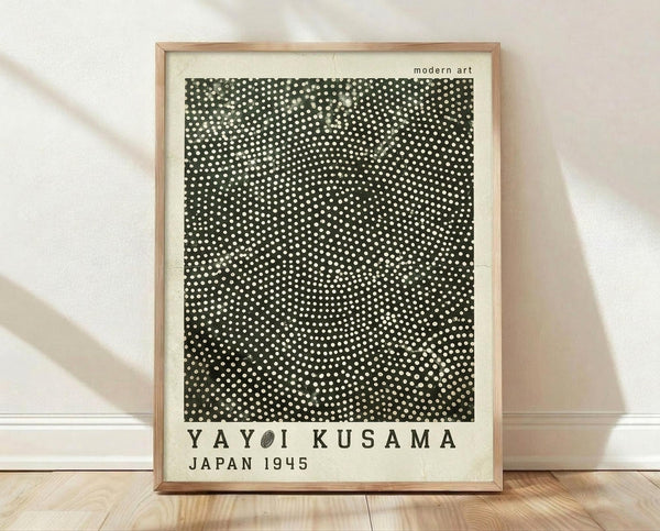 May include: Framed poster print with black background covered in white polka dots in varying sizes creating an organic pattern. Text reads "YAYOI KUSAMA JAPAN 1945" and "modern art" at top. Light wood frame sits on wooden floor against white textured wall with diagonal shadows.