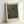May include: Framed modern art print featuring black background with white polka dots in varying sizes creating an optical illusion pattern. Light wood frame with white matting displays "YAYOI KUSAMA JAPAN 1945" text below artwork. Print shown leaning against white wall, casting soft shadow.