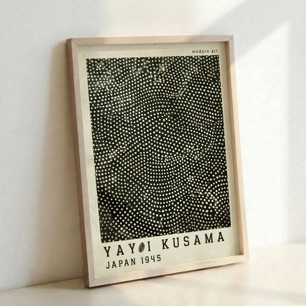 May include: Framed modern art print featuring black background with white polka dots in varying sizes creating an optical illusion pattern. Light wood frame with white matting displays "YAYOI KUSAMA JAPAN 1945" text below artwork. Print shown leaning against white wall, casting soft shadow.