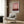 May include: Framed art print with pink and burgundy abstract watercolor design hanging on wood-paneled wall above beige upholstered armchair with geometric patterned pillow, brass floor lamp with white marble base, and dark round side table with open book in modern interior setting