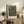 May include: Framed art print with black background and white polka dot pattern in light wood frame, displayed on rustic wooden sideboard alongside decorative pottery, books, and fern plant in neutral-toned living room setting with beige chair visible.