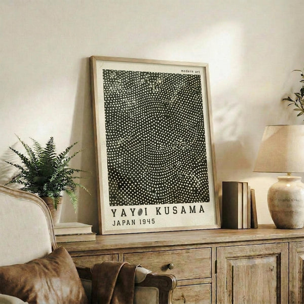 May include: Framed art print with black background and white polka dot pattern in light wood frame, displayed on rustic wooden sideboard alongside decorative pottery, books, and fern plant in neutral-toned living room setting with beige chair visible.