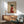 May include: Framed poster print displayed above wooden sideboard in modern living room setting. Artwork shows minimalist illustration of black greyhound head and neck wearing red knit sweater, holding white coffee mug, against beige background. Natural sunlight streams across white walls. Green upholstered chair with wooden arms and white table lamp visible in scene.