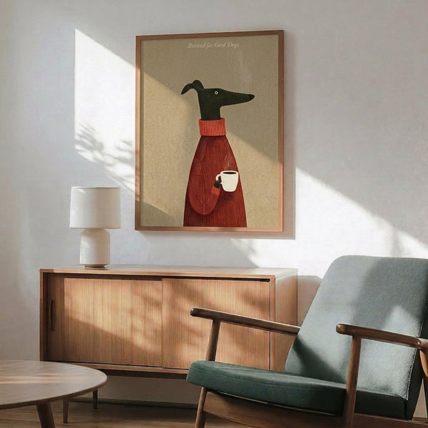 May include: Framed poster print displayed above wooden sideboard in modern living room setting. Artwork shows minimalist illustration of black greyhound head and neck wearing red knit sweater, holding white coffee mug, against beige background. Natural sunlight streams across white walls. Green upholstered chair with wooden arms and white table lamp visible in scene.