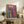 May include: Colorful framed art print with swirling psychedelic patterns in vibrant blues, purples, reds and yellows, featuring a figure looking toward flowing abstract forms and Japanese text at top. Light wood frame sits on rustic wooden sideboard next to fern plant, ceramic vase, and leather chair in neutral-toned room.