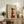 May include: Framed poster print featuring a stylized greyhound wearing a red ribbed sweater and holding a white coffee mug, displayed on a rustic wooden console table alongside books, ceramic vases, and a fern plant in a cozy neutral-toned living room setting with beige walls and warm lighting.