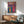 May include: Colorful surreal poster with Japanese characters displayed in modern living room setting. Artwork features vibrant flowing lines in rainbow colors depicting figure near waterfall landscape. Shown in light wood frame above walnut credenza with ceramic lamp. Mid-century green upholstered chair with wooden arms positioned nearby on hardwood floor against white wall with natural window light creating shadows.