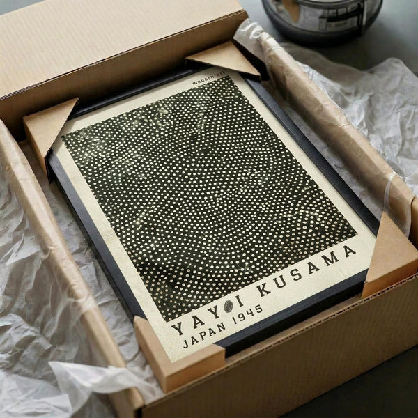 Framed artwork featuring a black background covered in white polka dots arranged in a gradient pattern, with text reading "YAYOI KUSAMA JAPAN 1945" at the bottom in cream colored lettering. The print is displayed in a black frame sitting in an open cardboard shipping box surrounded by white tissue paper, with wooden frame corners visible as packaging protection.