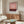 May include: Pink and burgundy abstract art print in white frame hanging above wooden sideboard in modern living room. White curved chair and glass coffee table with colorful surface visible. Warm natural lighting creates shadows on cream walls.