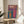 May include: Colorful abstract art print in light wood frame sitting on round wooden side table next to beige upholstered chair. Artwork features vibrant flowing lines in rainbow colors - purple, pink, orange, blue, green - with Japanese characters at top and silhouette figure. Bright sunlight streams across white wall creating geometric shadows. Green plant leaves visible in corner.