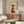 May include: Stylized illustration of a black greyhound wearing a red knit sweater and holding a white coffee cup, displayed in a thin frame above a walnut credenza in a modern living room with cream walls, white upholstered chair, and glass-top coffee table