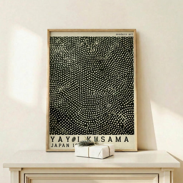 May include: Framed modern art poster featuring black background with dense white polka dot pattern creating optical illusion effect. Poster displays "YAYOI KUSAMA JAPAN 1945" text at bottom in black letters on cream background. Natural wood frame sits on light wooden surface against cream wall, with small wrapped white gift box and sprig of greenery placed in front.