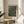 May include: Framed art print with black background covered in white polka dots, displayed on round wooden side table next to beige upholstered chair. Natural light streams across neutral walls, with plant leaves visible in corner. Print features text reading "YAYOI KUSAMA JAPAN 1945" below dotted pattern, housed in thin wooden frame with light matting.