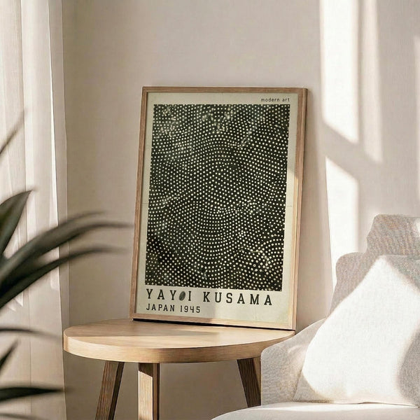May include: Framed art print with black background covered in white polka dots, displayed on round wooden side table next to beige upholstered chair. Natural light streams across neutral walls, with plant leaves visible in corner. Print features text reading "YAYOI KUSAMA JAPAN 1945" below dotted pattern, housed in thin wooden frame with light matting.