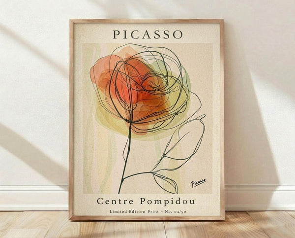 May include: Framed art print featuring abstract line drawing of a rose with orange and yellow watercolor washes on cream background, displayed on light wooden floor against white wall with natural shadows. The minimalist botanical artwork shows flowing black lines forming flower petals and stem with "PICASSO" and "Centre Pompidou" text, housed in natural wood frame.