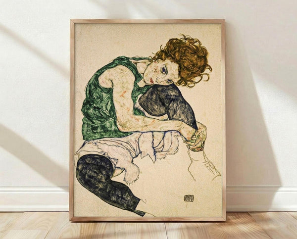 May include: Framed reproduction of an expressionist drawing showing a seated woman with curly reddish hair wearing a green sleeveless top and dark stockings, rendered in sketchy lines with muted colors on cream background. The wooden frame sits on light hardwood flooring against a white textured wall with soft natural lighting creating diagonal shadows.