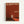 A minimalist art exhibition poster with a rust-colored background featuring a small silhouette of a person walking toward a bright white doorway. The figure casts a long shadow on what appears to be a reddish floor or ground. White text at the top reads "THE THRESHOLD PROJECT" and "WALKING INTO THE UNKNOWN" with exhibition details for Tate Modern, London. The poster has a light wooden frame and is photographed against a white wall.