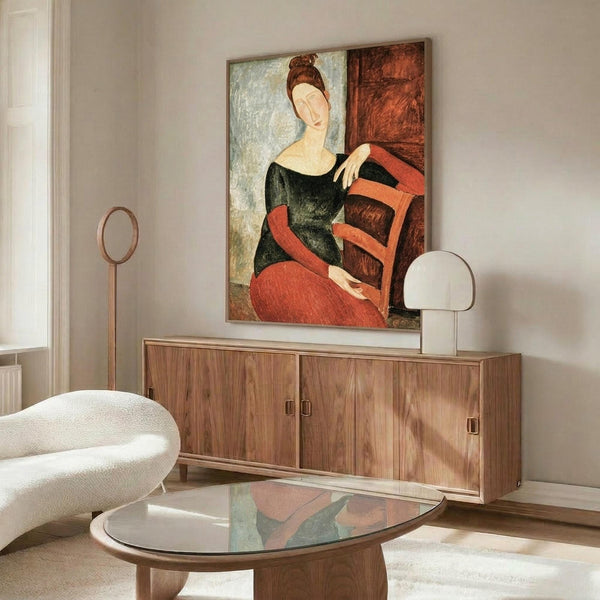 May include: A styled living room featuring a framed reproduction of Modigliani's portrait of Jeanne Hébuterne hanging above a wooden sideboard. The artwork shows a woman with elongated features in a dark green dress and coral-colored skirt, painted in the artist's distinctive style with warm earth tones. The modern interior includes a white curved chair, glass coffee table, and contemporary lighting.
