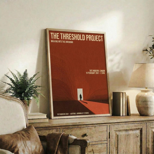 May include: Red minimalist art poster with white text reading "THE THRESHOLD PROJECT" and "WALKING INTO THE UNKNOWN" displayed in light wood frame on rustic wooden sideboard. Small silhouette figure visible in white doorway on red geometric background. Room setting includes fern plant, ceramic vase, books, and beige upholstered chair with brown leather pillow against cream wall.