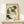 May include: Framed art print showing a woman with curly brown hair in a green sleeveless top and black stockings, sitting in a contemplative pose. The artwork features expressive line work and muted colors on cream background. Natural wood frame sits on light wooden surface next to small white wrapped gift against neutral wall.