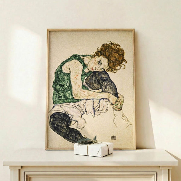May include: Framed art print showing a woman with curly brown hair in a green sleeveless top and black stockings, sitting in a contemplative pose. The artwork features expressive line work and muted colors on cream background. Natural wood frame sits on light wooden surface next to small white wrapped gift against neutral wall.