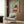 May include: Colorful retro poster featuring a anthropomorphic chicken character with yellow head and red comb, wearing polka dot outfit, positioned above orange turntable on teal background. Framed artwork hangs on wood-paneled wall above cream upholstered chair with geometric throw pillow. Modern brass floor lamp and dark side table with open book complete the contemporary living room setting.