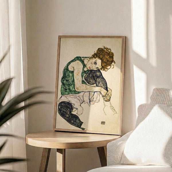May include: Framed reproduction of early 20th century expressionist artwork showing a seated woman with curly brown hair wearing a green blouse and dark stockings, displayed on a round wooden side table next to a beige upholstered chair with plant leaves visible in soft natural lighting