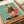 A whimsical illustration of a rooster wearing sunglasses and a black polka dot dress with coral accents, positioned as a DJ behind a vintage turntable. The rooster has a yellow head with red comb and wattle, set against a mint green background with a coral circle behind it. The turntable features black vinyl records with colorful striped elements in orange, coral, and cream tones. The artwork has a retro, mid-century modern aesthetic with textured paper appearance.