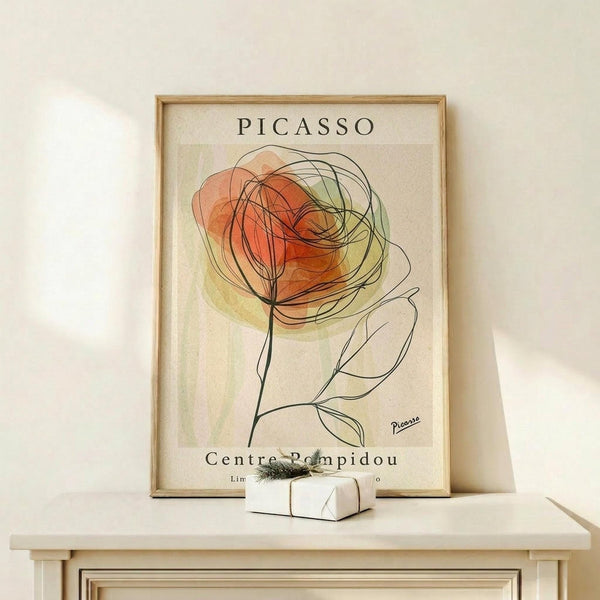 May include: Framed poster with abstract line drawing of a rose in orange and green watercolor tones, displaying "PICASSO" and "Centre Pompidou" text. Light wood frame sits on pale wooden shelf against cream wall, with small white wrapped gift box beside it. Minimalist modern home decor styling.