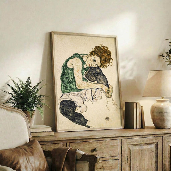 May include: Framed art print displaying a seated woman with curly brown hair wearing a green top and dark stockings, rendered in expressive line art style with muted colors on cream background. The wooden-framed piece sits on a rustic wooden sideboard alongside books, ceramic vases, and a fern plant, with a beige upholstered chair visible in the foreground of this neutral-toned living space.