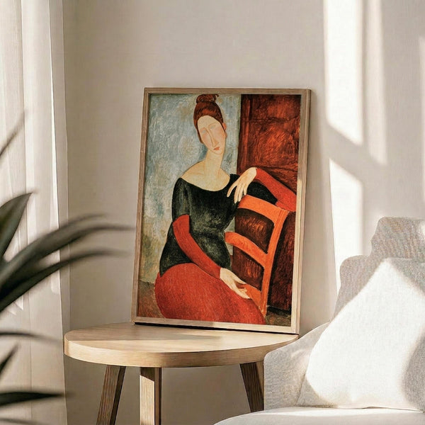 May include: Framed reproduction of Modigliani's painting featuring a woman with auburn hair in a black dress sitting on a red chair. The artwork shows the artist's characteristic elongated style with muted earth tones. Displayed on a round wooden side table next to a beige upholstered chair in a sunlit room with white walls and plant shadows.