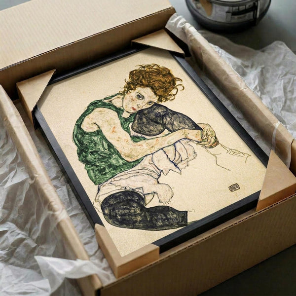 May include: Framed reproduction of an expressionist artwork showing a seated woman with curly reddish hair wearing a green sleeveless top and white skirt with black stockings. The drawing features bold outlines and muted earth tones on cream paper. Black frame sits in shipping box with protective tissue paper and cardboard tubes nearby on white surface.