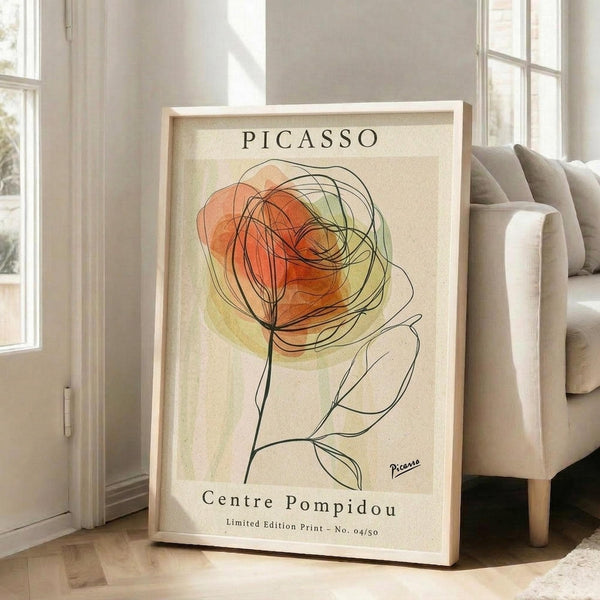 May include: Framed poster featuring a minimalist line drawing of a rose in Picasso's style, with orange and green watercolor washes on cream background, displayed on wooden floor next to beige sofa near bright windows in modern living room setting