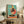 May include: Framed poster with turquoise background showing a stylized rooster in polka dot vest standing on a vinyl record turntable. The chicken has yellow body, red comb, and is positioned against a coral circle. Artwork displayed on rustic wooden sideboard with books, ceramic vase, and fern plant beside beige armchair in modern living room setting.