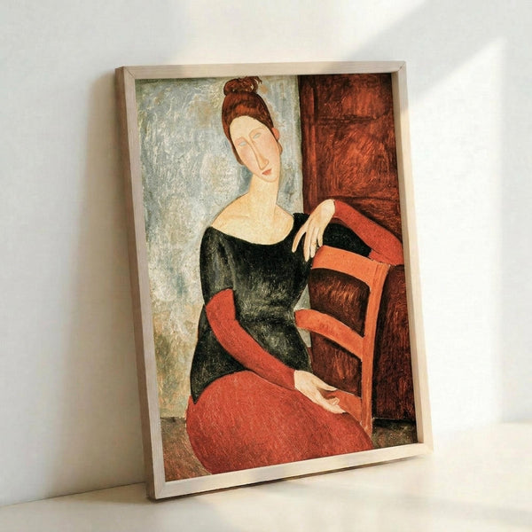 May include: A framed art print leaning against a white wall showing a woman with auburn hair in a bun, wearing a black top and coral-red skirt, seated in a wooden chair. The painting features elongated forms and muted colors typical of early 20th century modernist style, with a pale blue-green background. The print is displayed in a simple light wood frame.