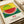A colorful pointillism-style watermelon illustration on white paper in an open book or portfolio. The watermelon slice shows green striped rind and red flesh with black seeds against a yellow dotted background. Below the artwork are Japanese characters and English text about courage. The paper has a cream-colored tone and sits on what appears to be a white surface with rolled papers nearby.