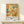 May include: Framed Paul Klee exhibition poster displaying colorful abstract artwork with small human figures in warm oranges, teals, and earth tones against textured background. Wooden frame rests on light wood shelf beside wrapped gift with sprig of greenery. White wall background with soft natural lighting creates minimalist display setting.