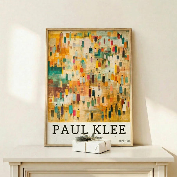 May include: Framed Paul Klee exhibition poster displaying colorful abstract artwork with small human figures in warm oranges, teals, and earth tones against textured background. Wooden frame rests on light wood shelf beside wrapped gift with sprig of greenery. White wall background with soft natural lighting creates minimalist display setting.