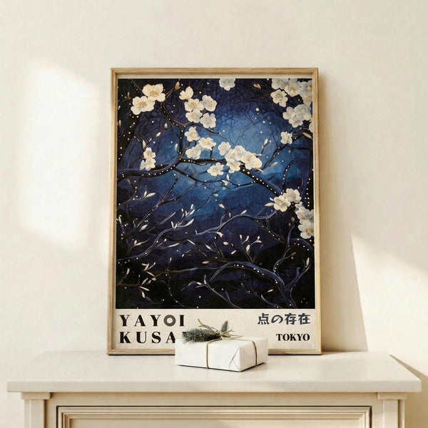 May include: Framed art print displayed on light wood shelf showing white cherry blossoms against deep blue night sky with scattered dots, styled as Japanese-inspired artwork. Text reads "YAYOI KUSAMA" and Japanese characters with "TOKYO" below. Small wrapped gift box with evergreen sprig sits beside the natural wood frame on minimalist shelf against cream wall.