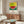 May include: Colorful watermelon art print with Japanese text displayed above wooden credenza in modern living room. The pointillism-style artwork shows a green watermelon and red slice against yellow and blue background, framed in light wood. Room features cream walls, white curved chair, oval glass coffee table, and brass floor lamp.